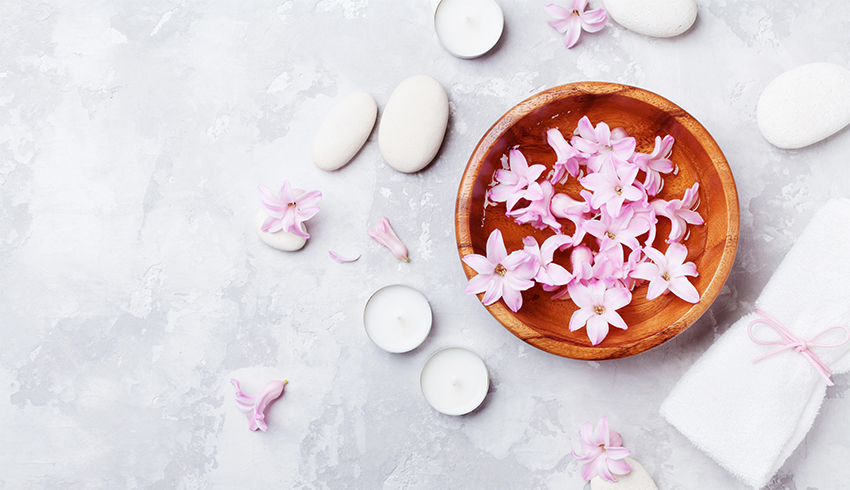 Neroli flower blossoms sit in a wooden bowl filled with clear liquid and surrounded by tea lights, massage stones, a white folded towel and more blossoms.